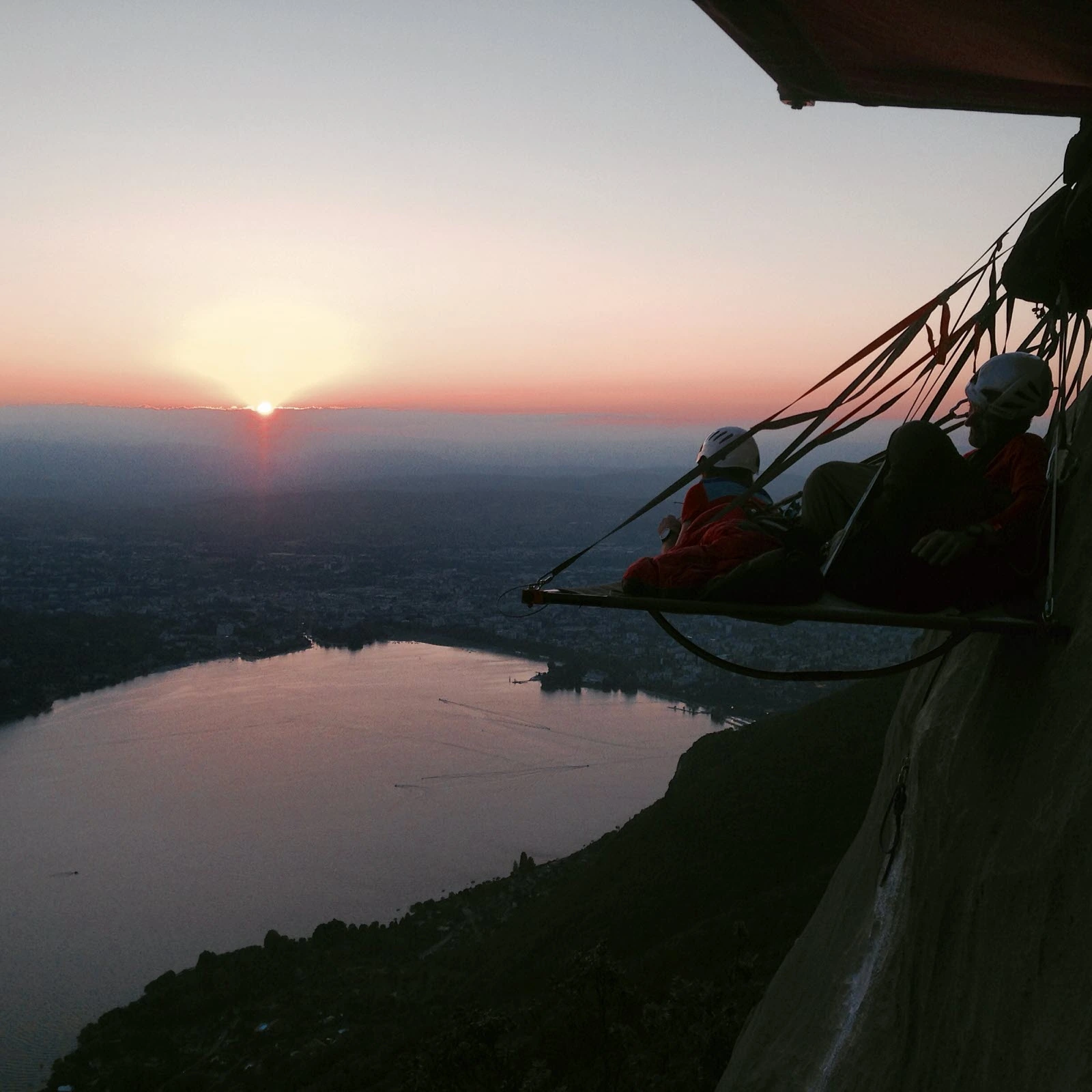 Deux grimpeurs sur un portaledge suspendu à la paroi, coucher de soleil sur le lac et la vallée en contrebas