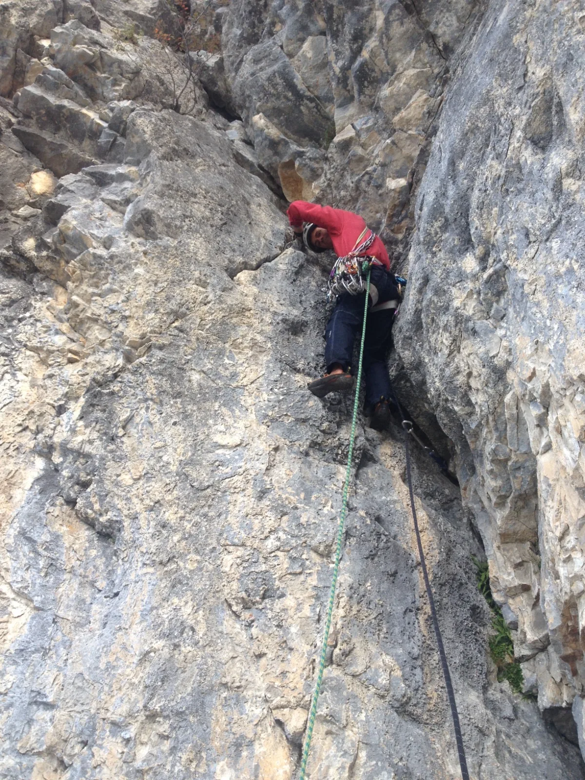 Grimpeur en veste rouge dans un dièdre de calcaire gris et ocre, vu depuis le bas, corde qui descend vers l'assureur