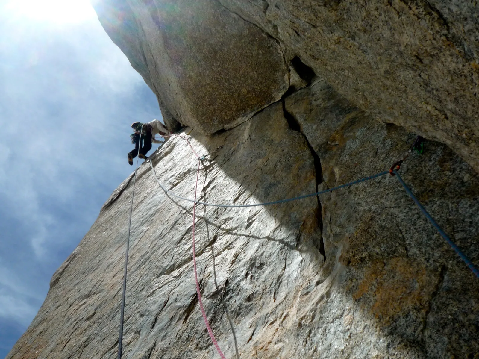 Grimpeur en haut d'une dalle de granite fissurée, la corde tendue en diagonale sous lui, ciel nuageux