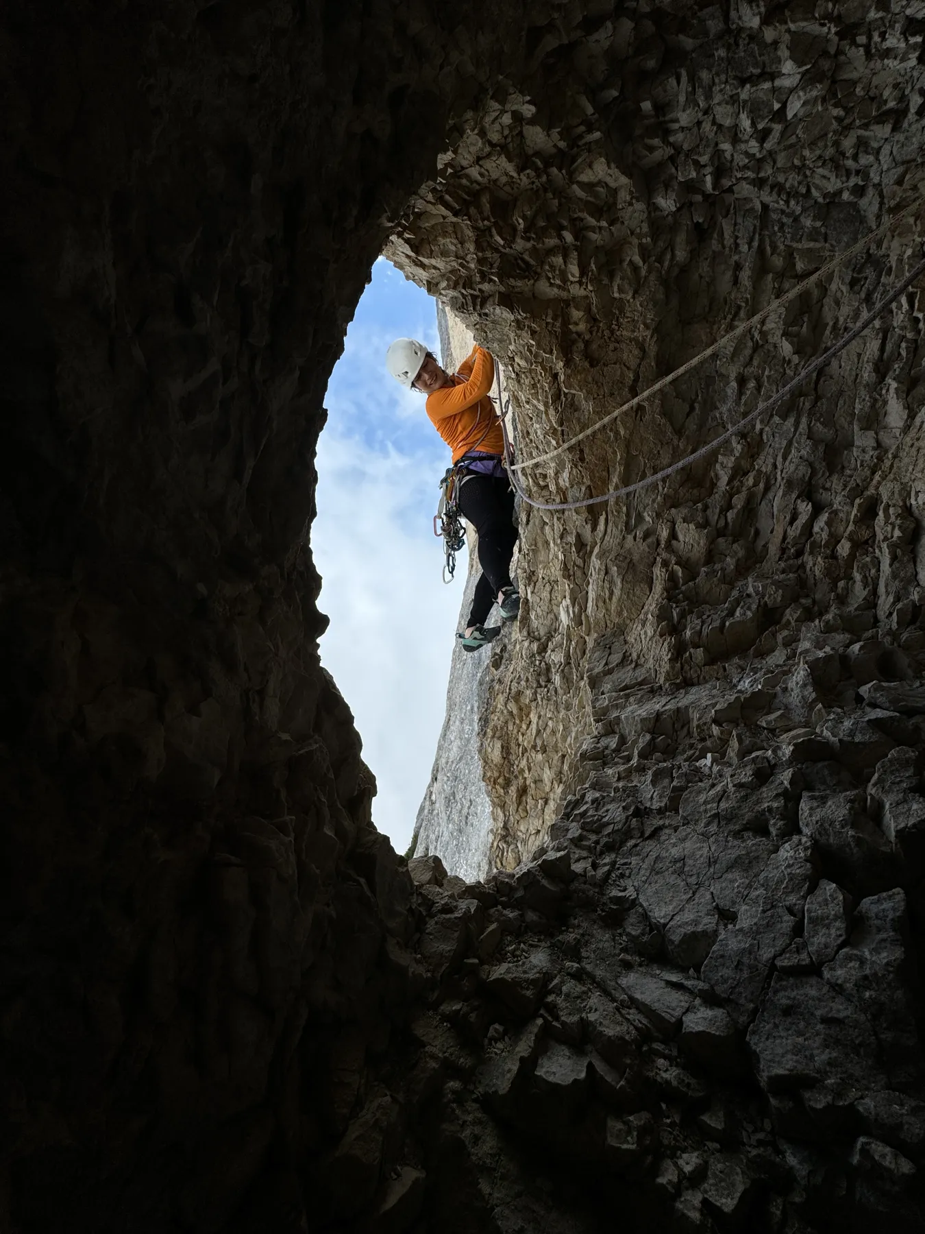 Grimpeuse franchissant l'ouverture lumineuse d'une grotte en paroi, ciel bleu derrière